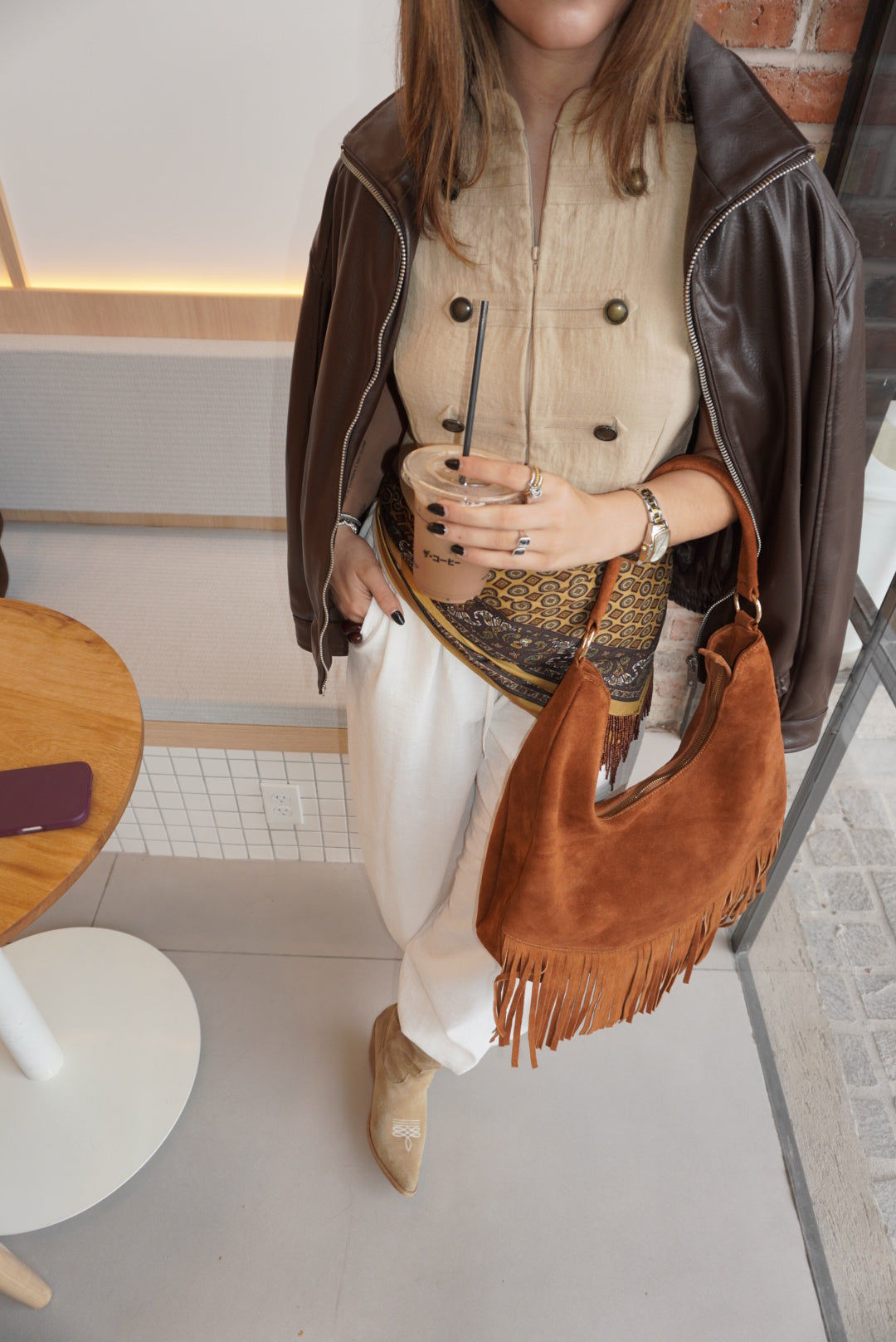 A woman wearing a brown faux-leather jacket, cream double-breasted vest, white trousers, and tan ankle boots, carrying a brown suede fringe shoulder bag while holding a drink in a modern indoor setting.