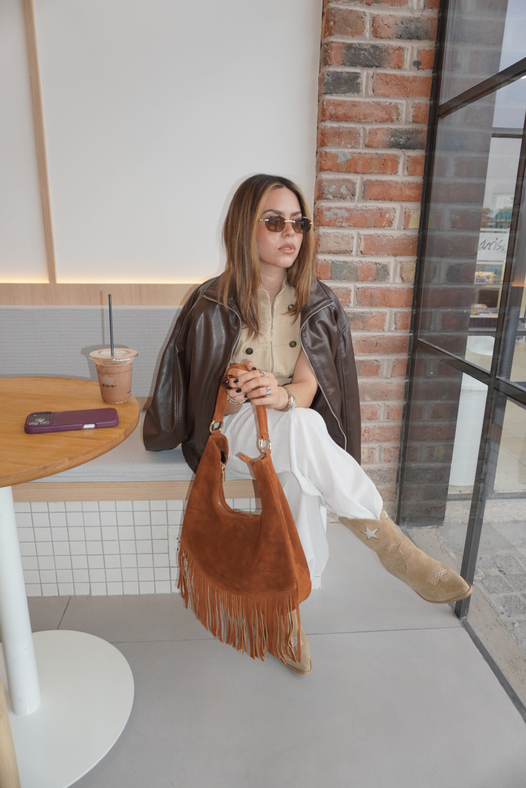 A woman wearing sunglasses, a brown leather jacket, a beige top, and white pants sits in a cafe setting, holding a large brown suede fringe shoulder bag.